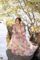 A woman in a floral dress sitting on a rock by the water.