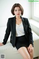 A woman in a black blazer and skirt sitting on a white couch.