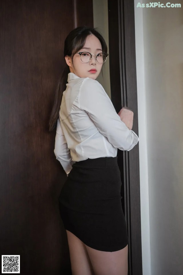 A woman in a white shirt and black skirt leaning against a wall.