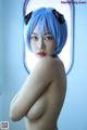 A woman with blue hair is posing naked in front of a window.