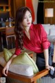 A woman in a red shirt and black skirt leaning on a chair.